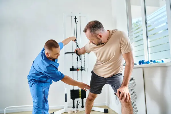 Medico esperto in uniforme blu che aiuta l'uomo a lavorare sulla macchina da allenamento nel centro di cinesiologia — Foto stock