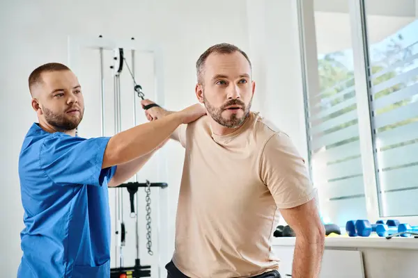 Young physician in blue uniform helping man working out on training machine in kinesiology center — Stock Photo