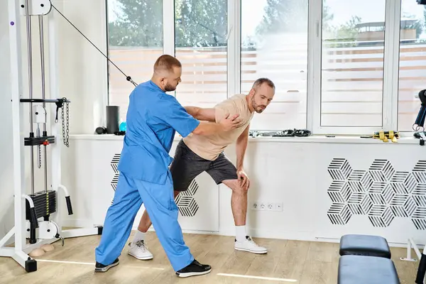 Professional instructor in blue uniform helping man during recovery training in kinesiology center — Stock Photo