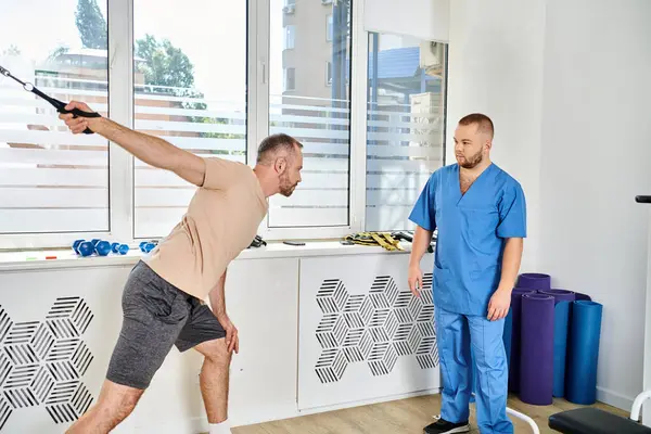Mann in Sportbekleidung trainiert an Trainingsgerät in der Nähe eines erfahrenen Rehabilitologen im Kinesio-Zentrum — Stockfoto