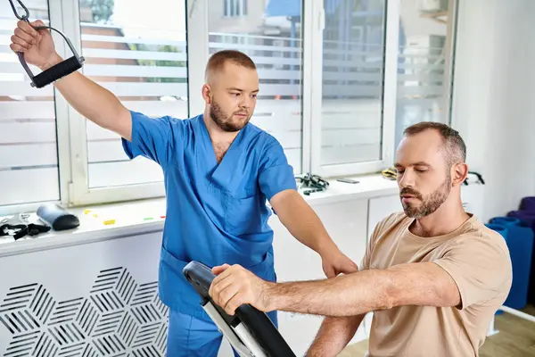 Bearded man in sportswear training on exercise machine near young physician in kinesio center — Stock Photo