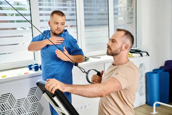 Junge Rehabilitologin in blauer Uniform assistiert Mann beim Aufwachtraining im Kinesio-Zentrum — Stockfoto