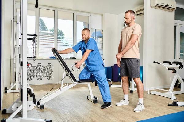 Skilled instructor in blue uniform showing how to train on exercise machine in kinesiology center — Stock Photo