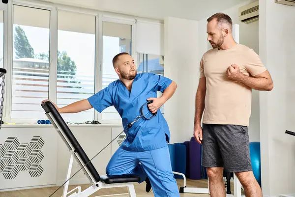 Молодой врач в синей форме инструктирует человека, как тренироваться на тренажере в кинезиологическом центре — стоковое фото