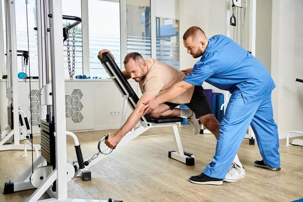 Rehabilitólogo en uniforme azul enseñando al hombre cómo entrenar en la máquina de ejercicio en el centro de kinesiología - foto de stock