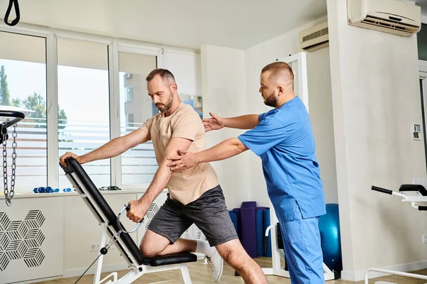 Professional instructor supporting man in sportswear during recovery training on exercise machine — Stock Photo