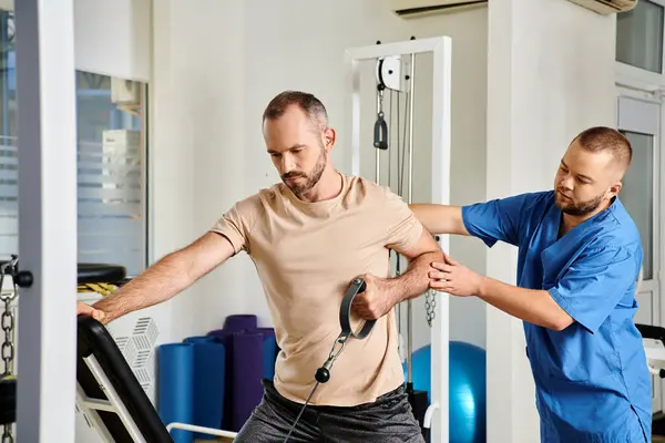 Young skilled physician supporting man in sportswear during recovery training on exercise machine — Stock Photo