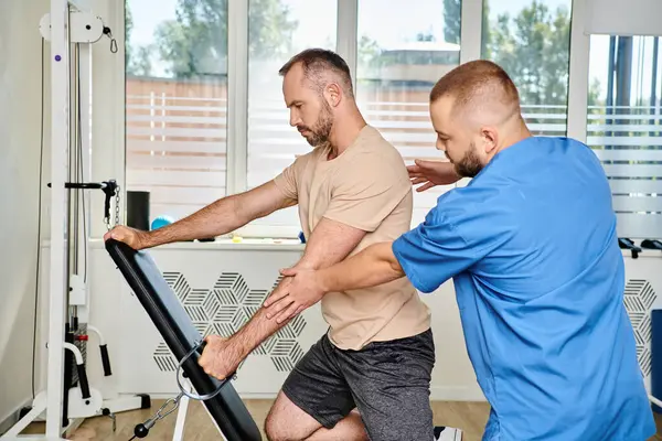 Опытный физиотерапевт помогает человеку в спортивной одежде во время восстановительных тренировок на тренажере — стоковое фото