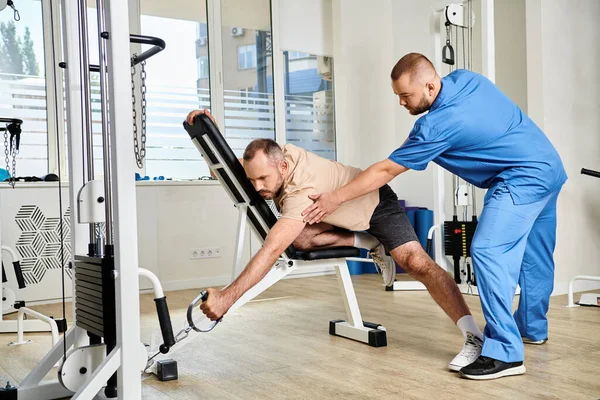 Medico in uniforme blu che aiuta l'uomo durante l'allenamento di recupero sulla macchina da ginnastica nel centro di Kinesio — Foto stock