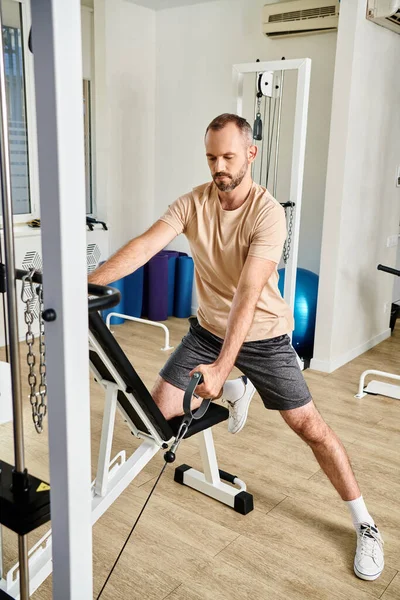 Bearded man in sportswear working out on exercise machine in modern gym of kinesio center, recovery — Stock Photo