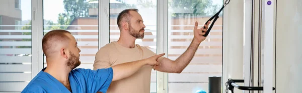 Médico qualificado em uniforme azul instruindo o homem no ginásio do centro de kinesio, bandeira horizontal — Fotografia de Stock