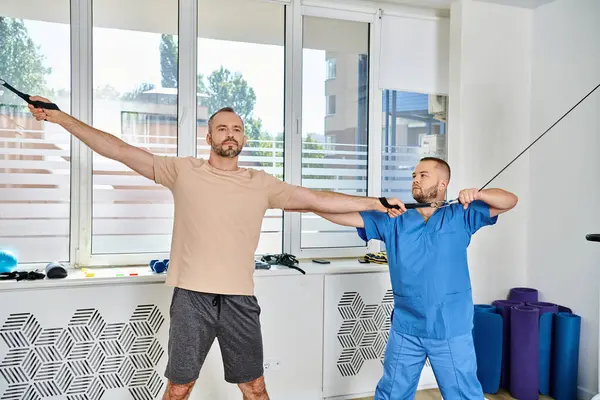 Médecin expérimenté en uniforme bleu soutenant la formation de l'homme sur la machine d'exercice dans le centre de kinesio — Photo de stock