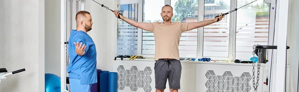 Recovery specialist assisting man training on exercise machine in modern kinesio center, banner — Stock Photo