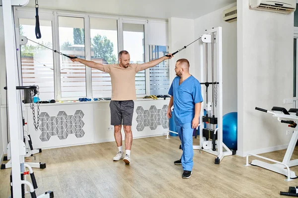 Fisioterapeuta experto en uniforme azul que ayuda al hombre a entrenar en la máquina de ejercicio en el centro kinesio - foto de stock