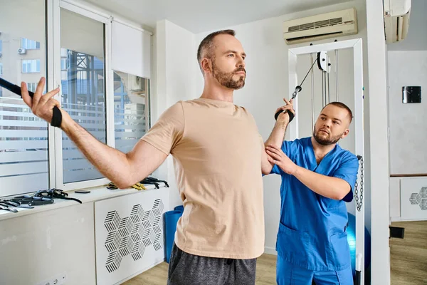 Skilled physician helping man in sportswear training on exercise machine in modern kinesio center — Stock Photo