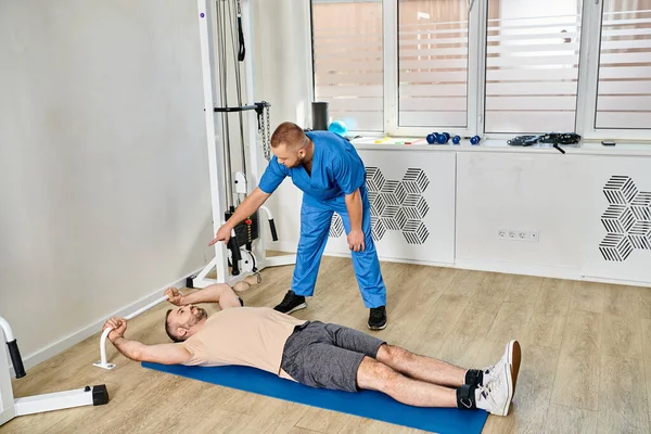 Professioneller Arzt, der dem Mann beim Sportbekleidungstraining auf dem Trainingsgerät im Kinesio-Zentrum hilft — Stockfoto
