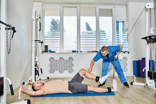 Rehabilitólogo experimentado que apoya el entrenamiento del hombre en la máquina del ejercicio en el gimnasio del centro de kinesio - foto de stock