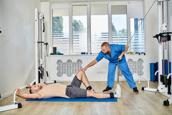 Квалифицированный физиотерапевт, поддерживающий тренировку человека на тренажере в тренажерном зале кинезио-центра — стоковое фото