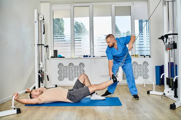 Молодий лікар у блакитній формі допомагає чоловікові тренуватися на тренажерах в тренажерному залі центру Кінезіо — стокове фото