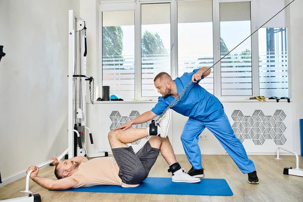 Кваліфікований лікар у блакитній формі допомагає чоловікові тренуватися на тренажерному автоматі в тренажерному залі центру Кінезіо — стокове фото