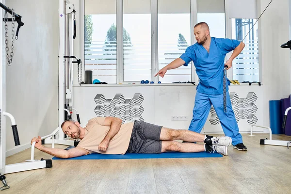 Лікар у блакитній формі вказує і навчає чоловіка під час тренувань з відновлення в центрі кінезіо — стокове фото