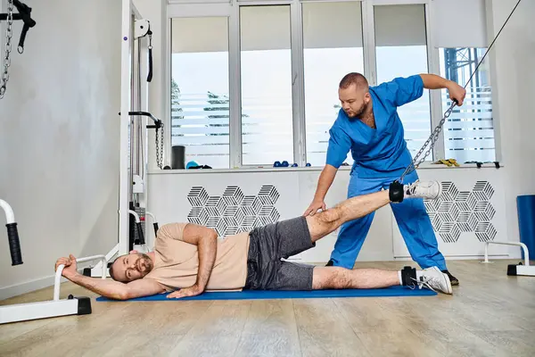 Qualifizierter Rehabilitologe in blauer Uniform, der dem Mann beim Rehabilitationstraining im Kinesiozentrum assistiert — Stockfoto