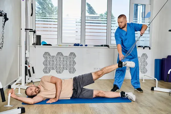 Instructor experimentado en uniforme azul ayudando al hombre durante el entrenamiento de recuperación en el centro kinesio - foto de stock
