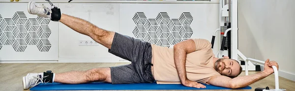 Bearded man lying down on fitness mat and training on exercise machine in kinesio center, banner — Stock Photo