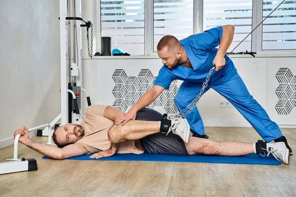 Young skilled rehabilitologist assisting man working out during recovery in modern kinesio center — Stock Photo
