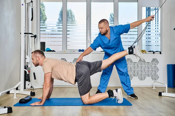 Qualifizierte Physiotherapeutin unterstützt den Mann beim Training während der Erholungsphase in der Turnhalle des Kinesio-Zentrums — Stockfoto
