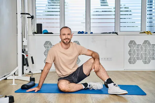 Homem bonito feliz em sportswear sentado no tapete de fitness perto de máquina de exercício no centro de kinesio — Fotografia de Stock