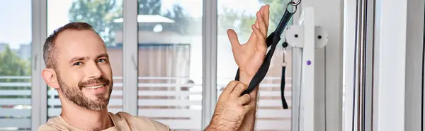 Joyful bearded man looking at camera near modern exercise machine in kinesio center, banner — Stock Photo