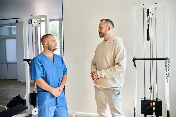 Бородатый мужчина и профессиональный реабилитолог в синей форме разговаривают в современном центре кинезио — стоковое фото