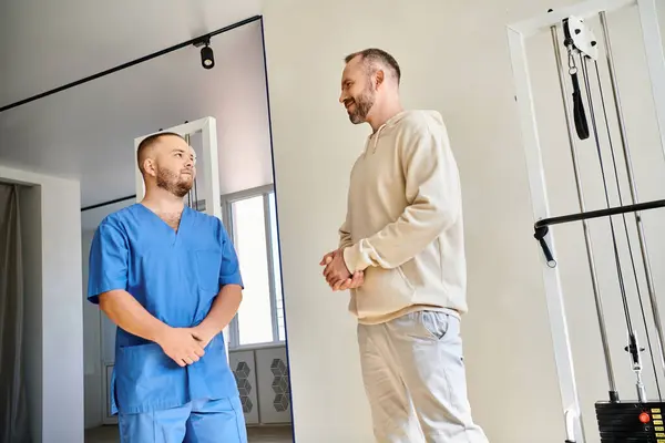 Reabilitologista experiente em uniforme azul conversando com paciente masculino no moderno centro de cinesio — Fotografia de Stock