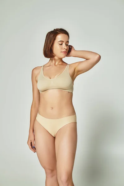 Cuerpo positivo, mujer joven bonita con el pelo corto de pie en ropa interior beige sobre fondo gris - foto de stock