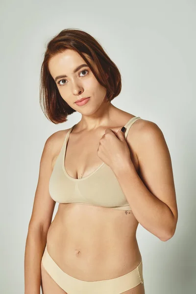 Self-assured and young woman in comfortable beige lingerie looking at camera on grey background — Stock Photo