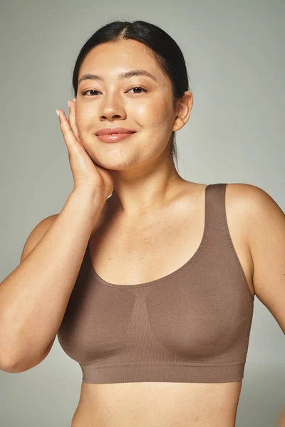 Jolie femme asiatique en haut brun touchant son visage sur fond gris, soins de la peau et corps positif — Photo de stock