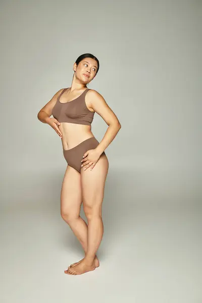 Pleine longueur de jolie jeune femme asiatique en sous-vêtements beige posant sur fond gris neutre — Photo de stock