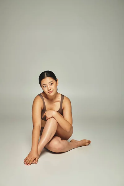Longitud completa de la joven mujer asiática en ropa interior sentado con las piernas cruzadas sobre fondo gris - foto de stock