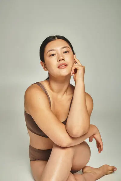 Brunette jeune femme asiatique en sous-vêtements assis avec les jambes croisées sur fond gris, corps positif — Photo de stock