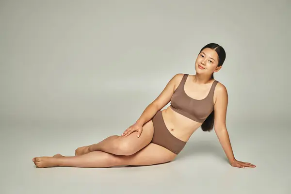 Полная длина азиатской женщины в нижнем белье сидя с босыми ногами на сером фоне, тело положительное — стоковое фото