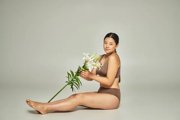 Young and brunette asian woman in underwear holding flowers and sitting on grey background, lilies — Stock Photo