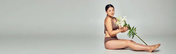 Young and brunette asian woman in underwear holding flowers and sitting on grey background, banner — Stock Photo