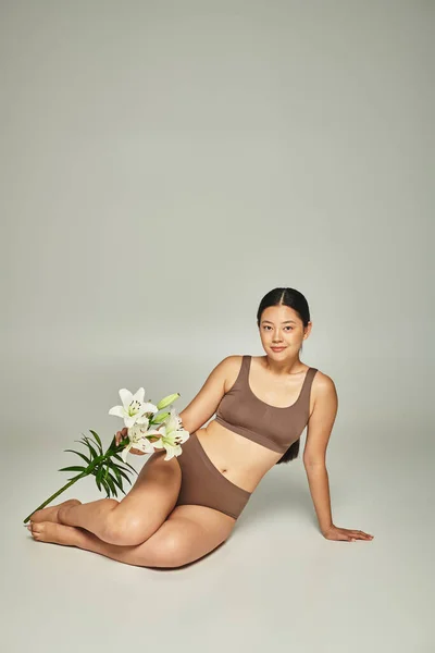 Brunette asian woman in pastel underwear holding flowers and sitting on grey background, lilies — Stock Photo