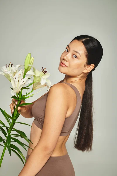 Brunette asian woman in pastel underwear holding flowers and standing on grey background, lilies — Stock Photo