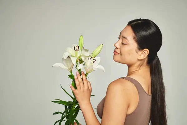 Heureux jeune asiatique femme en pastel sous-vêtements à la recherche de fleurs sur fond gris, lis — Photo de stock