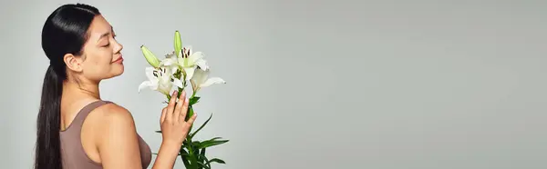 Heureux jeune asiatique femme en pastel sous-vêtements à la recherche de fleurs sur fond gris, bannière — Photo de stock