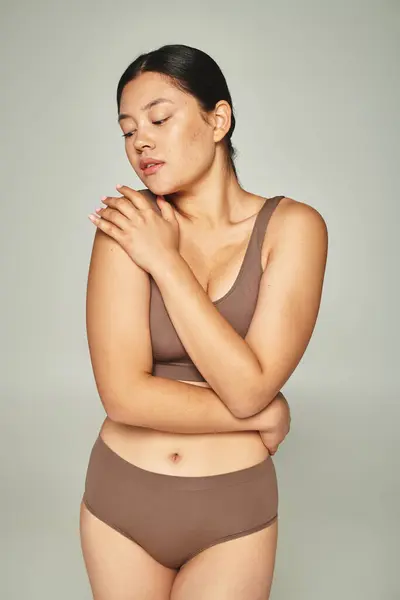 Donna asiatica in biancheria intima che copre il corpo abbracciandosi su sfondo grigio, femminile — Foto stock