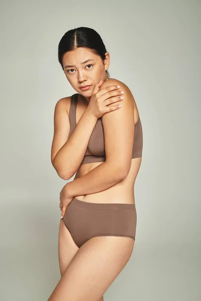 Joven asiático mujer en ropa interior cubierta cuerpo mientras abraza a sí misma en gris telón de fondo, cuerpo vergonzoso - foto de stock