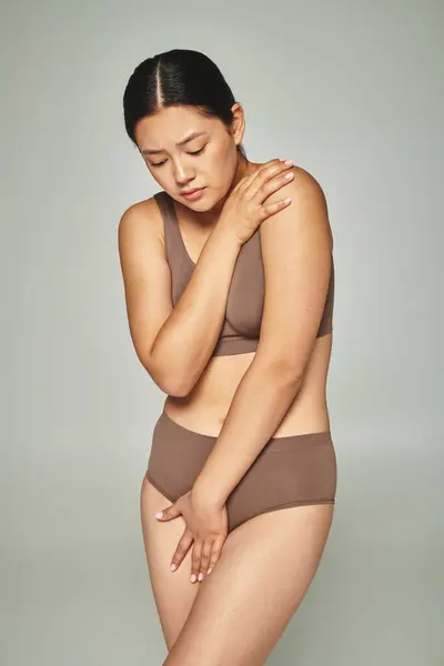 Sad asian woman in underwear covering body while embracing herself on grey backdrop, body shaming — Stock Photo